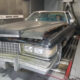 Dusty vintage Cadillac with hood open and front-end damage in a workshop; a person sits in the driver's seat.