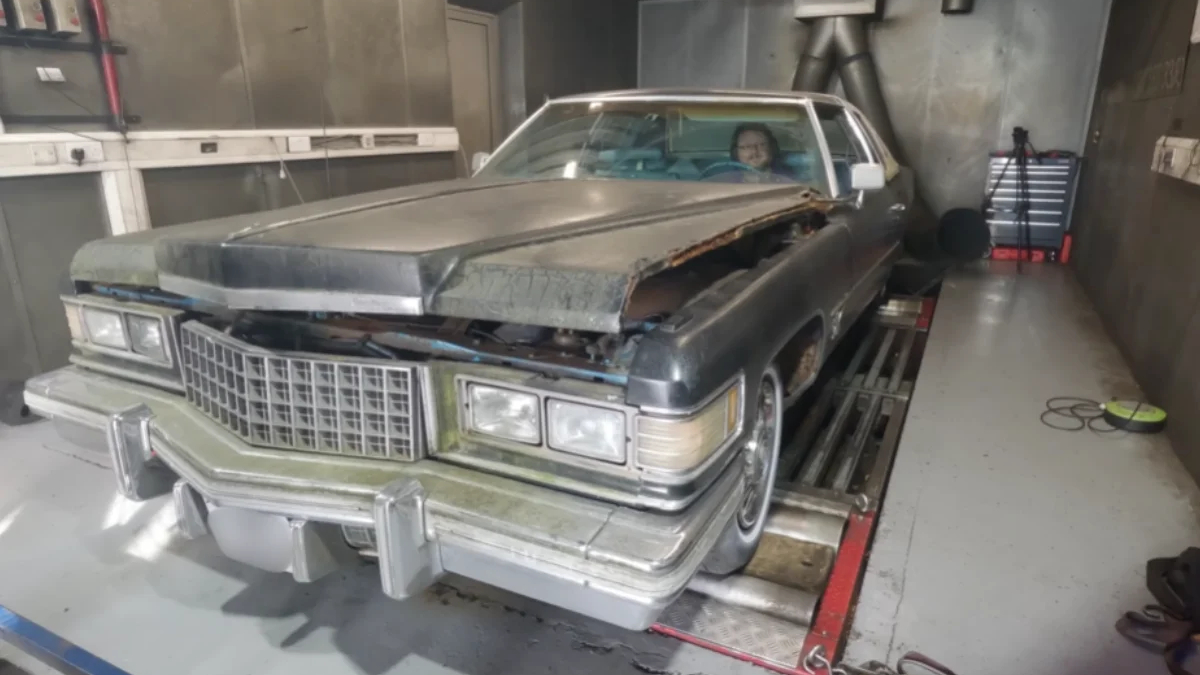 Dusty vintage Cadillac with hood open and front-end damage in a workshop; a person sits in the driver's seat.