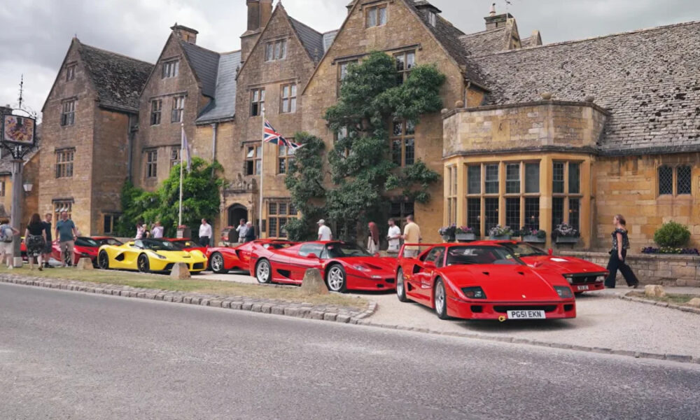 Row of red and yellow sports cars parked along a historic stone building as people admire them in a town square near Union Jack flags.