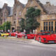 Row of red and yellow sports cars parked along a historic stone building as people admire them in a town square near Union Jack flags.