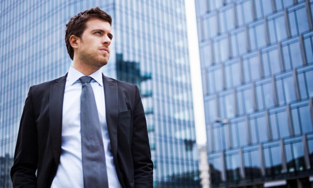 Young man in a dark suit and gray tie standing outdoors near glass office buildings in a cityscape.