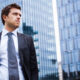 Young man in a dark suit and gray tie standing outdoors near glass office buildings in a cityscape.