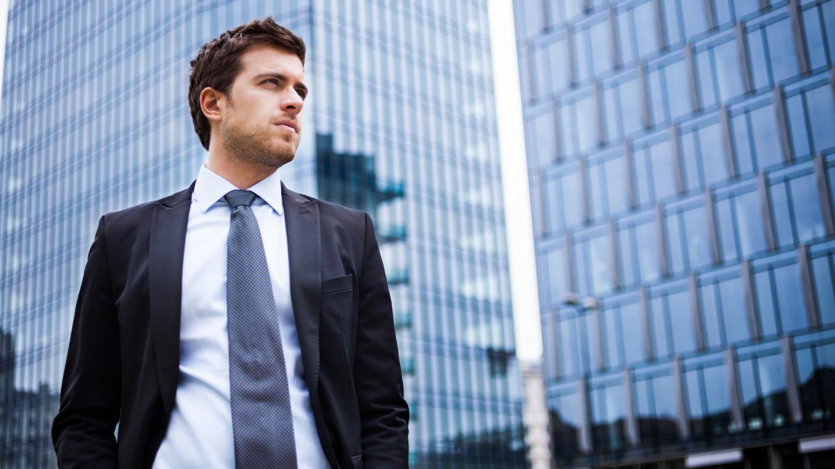 Young man in a dark suit and gray tie standing outdoors near glass office buildings in a cityscape.