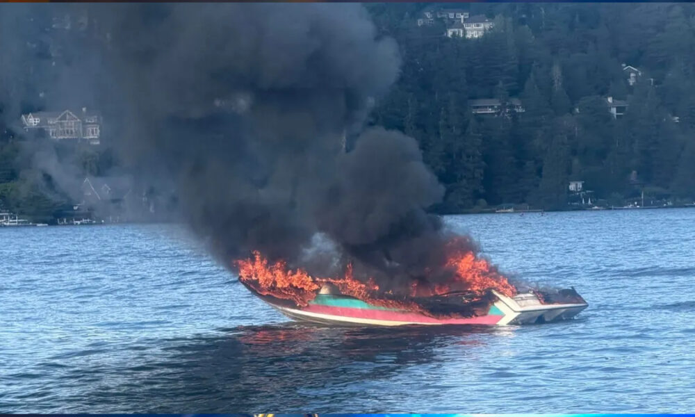 A colorful speedboat on a lake is engulfed in flames with thick black smoke rising above the water to the forested shore behind it.