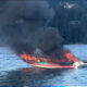 A colorful speedboat on a lake is engulfed in flames with thick black smoke rising above the water to the forested shore behind it.