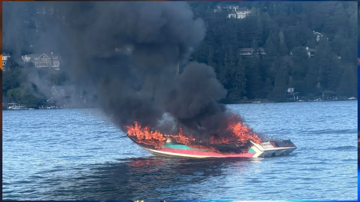 A colorful speedboat on a lake is engulfed in flames with thick black smoke rising above the water to the forested shore behind it.