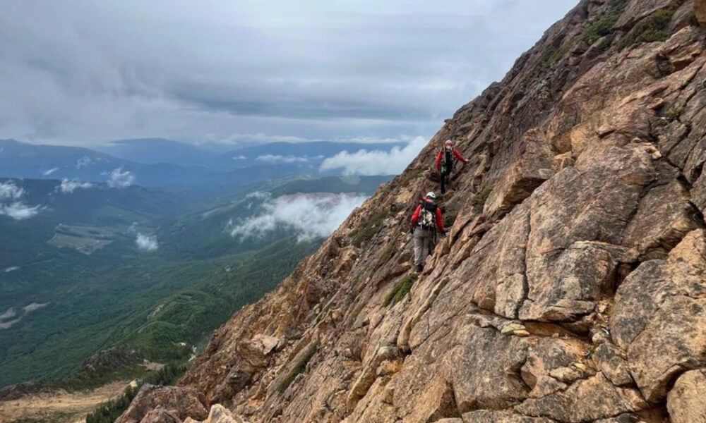 Two hikers in red jackets carefully traverse a steep rocky mountainside with backpacks, high above a valley.