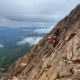 Two hikers in red jackets carefully traverse a steep rocky mountainside with backpacks, high above a valley.