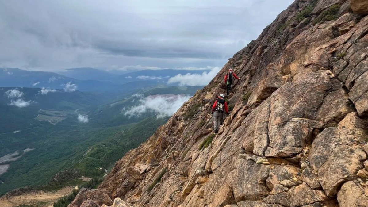Two hikers in red jackets carefully traverse a steep rocky mountainside with backpacks, high above a valley.