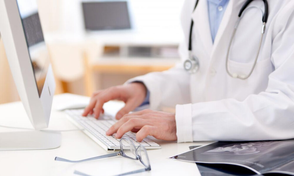 Doctor in a white coat typing on a computer keyboard at a desk, stethoscope around the neck.