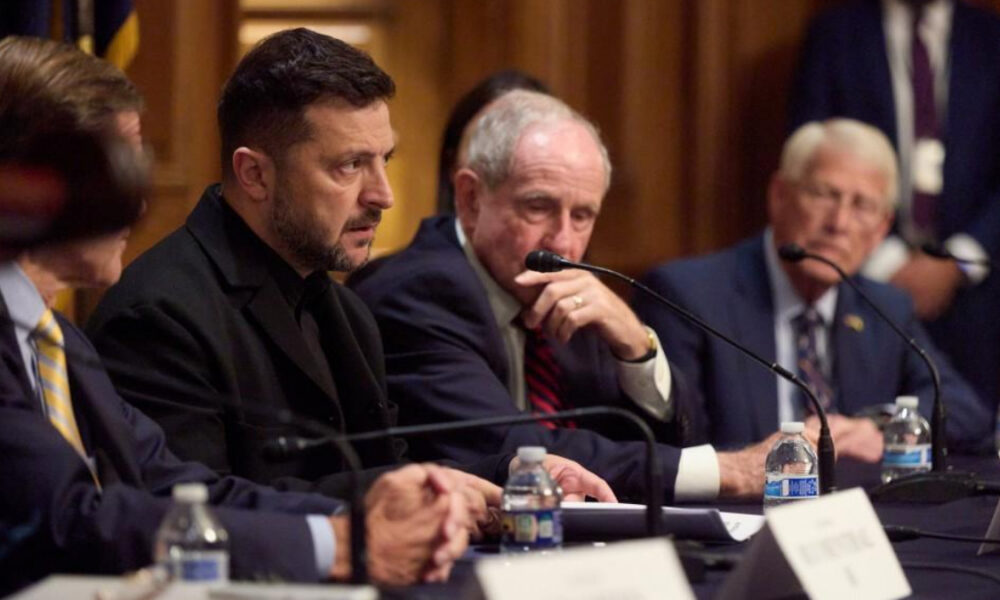 Panel of suited men at a formal meeting with microphones and water bottles on the table.