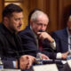 Panel of suited men at a formal meeting with microphones and water bottles on the table.