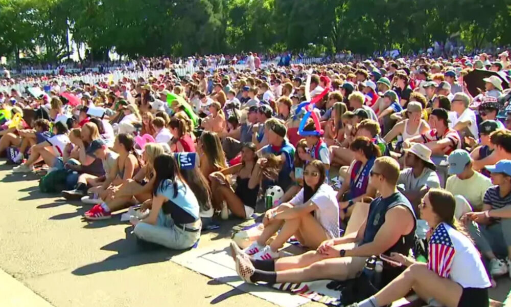 Large crowd of people seated on the ground at an outdoor event on a sunny day, many wearing hats and sunglasses in a park setting.