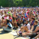 Large crowd of people seated on the ground at an outdoor event on a sunny day, many wearing hats and sunglasses in a park setting.