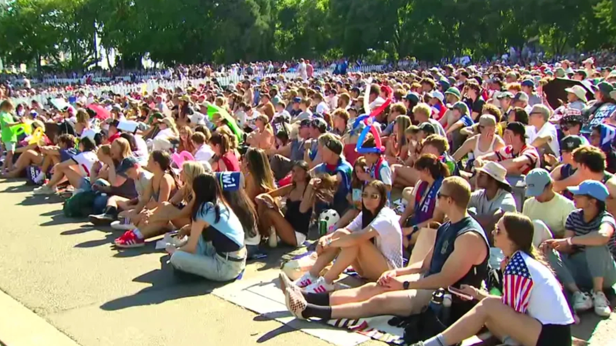 Large crowd of people seated on the ground at an outdoor event on a sunny day, many wearing hats and sunglasses in a park setting.