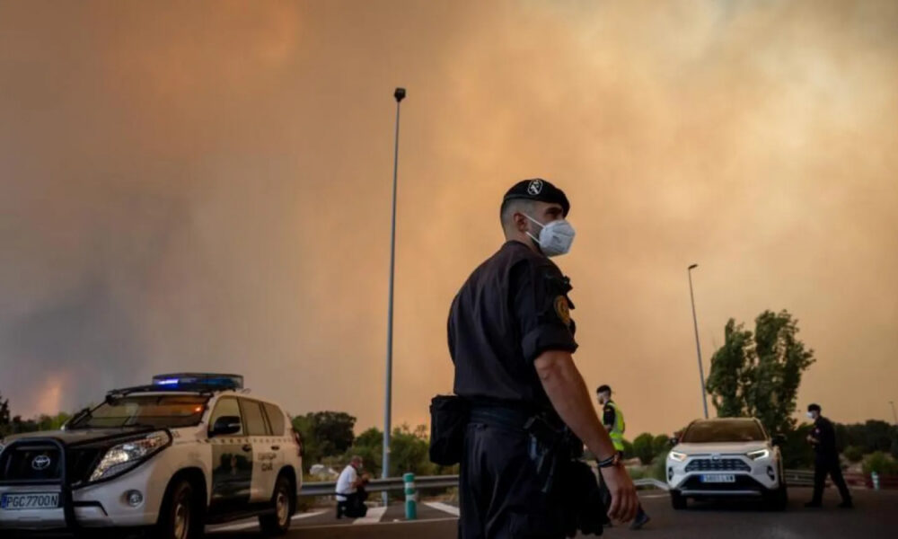 Police officer wearing a mask stands on a smoky street with an orange sky, patrol cars in the background and another officer nearby.