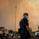 Police officer wearing a mask stands on a smoky street with an orange sky, patrol cars in the background and another officer nearby.