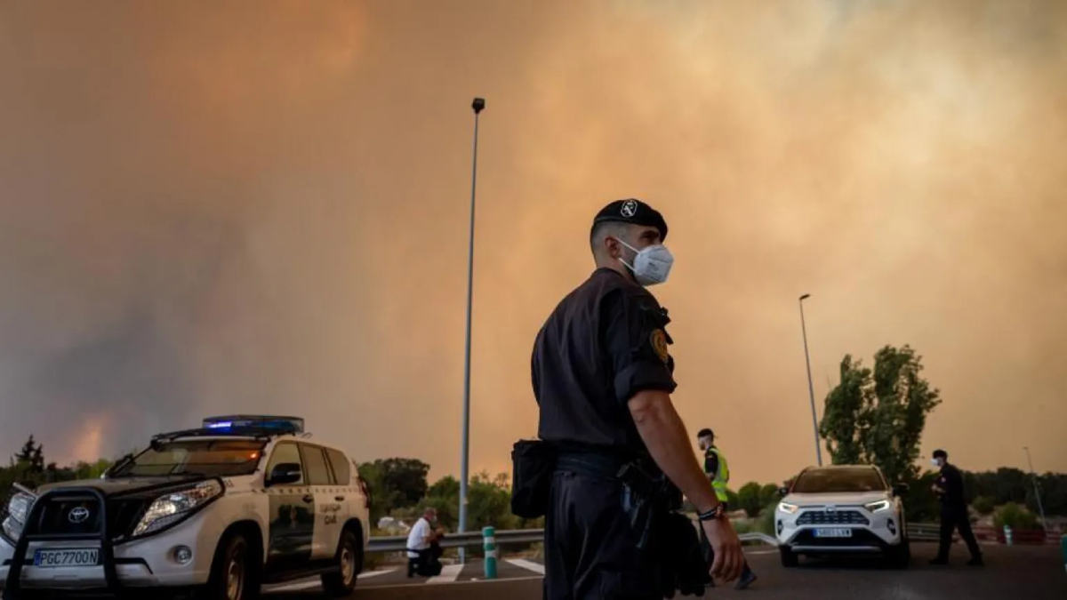 Police officer wearing a mask stands on a smoky street with an orange sky, patrol cars in the background and another officer nearby.