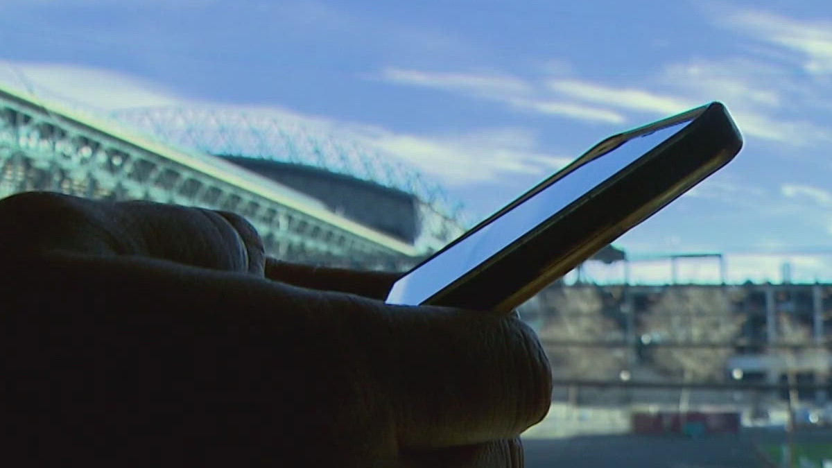 Close-up of a person holding a smartphone outdoors with a stadium and blue sky in the background.