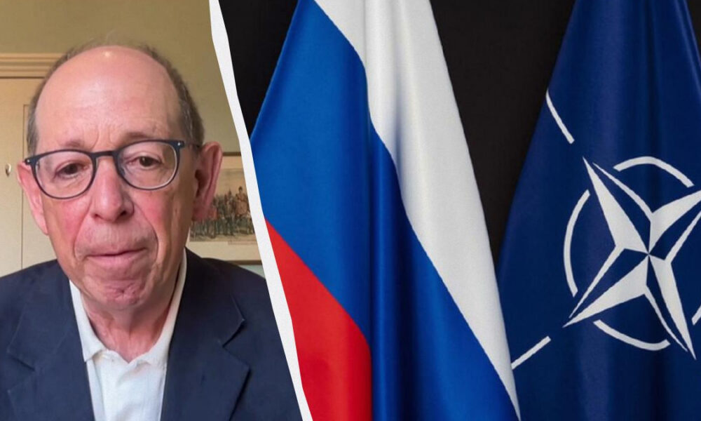 Man in a suit and glasses on a video call, with Russian and NATO flags visible on the right side of the image.