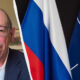 Man in a suit and glasses on a video call, with Russian and NATO flags visible on the right side of the image.