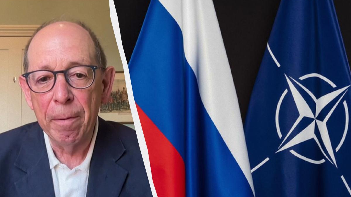 Man in a suit and glasses on a video call, with Russian and NATO flags visible on the right side of the image.