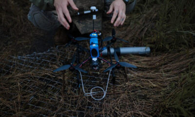 Quadcopter drone with a blue body sits on pine needles as a person adjusts it outdoors in a forest clearing.
