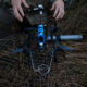 Quadcopter drone with a blue body sits on pine needles as a person adjusts it outdoors in a forest clearing.