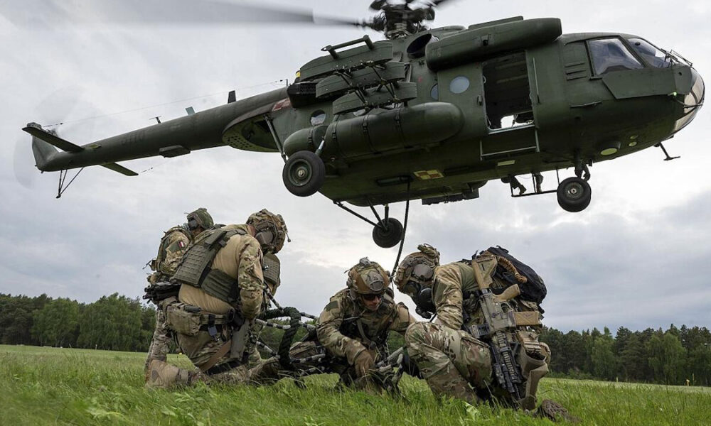 CH-47 Chinook helicopter hovering over soldiers in camouflage gear on a grassy field, preparing equipment.