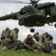 CH-47 Chinook helicopter hovering over soldiers in camouflage gear on a grassy field, preparing equipment.