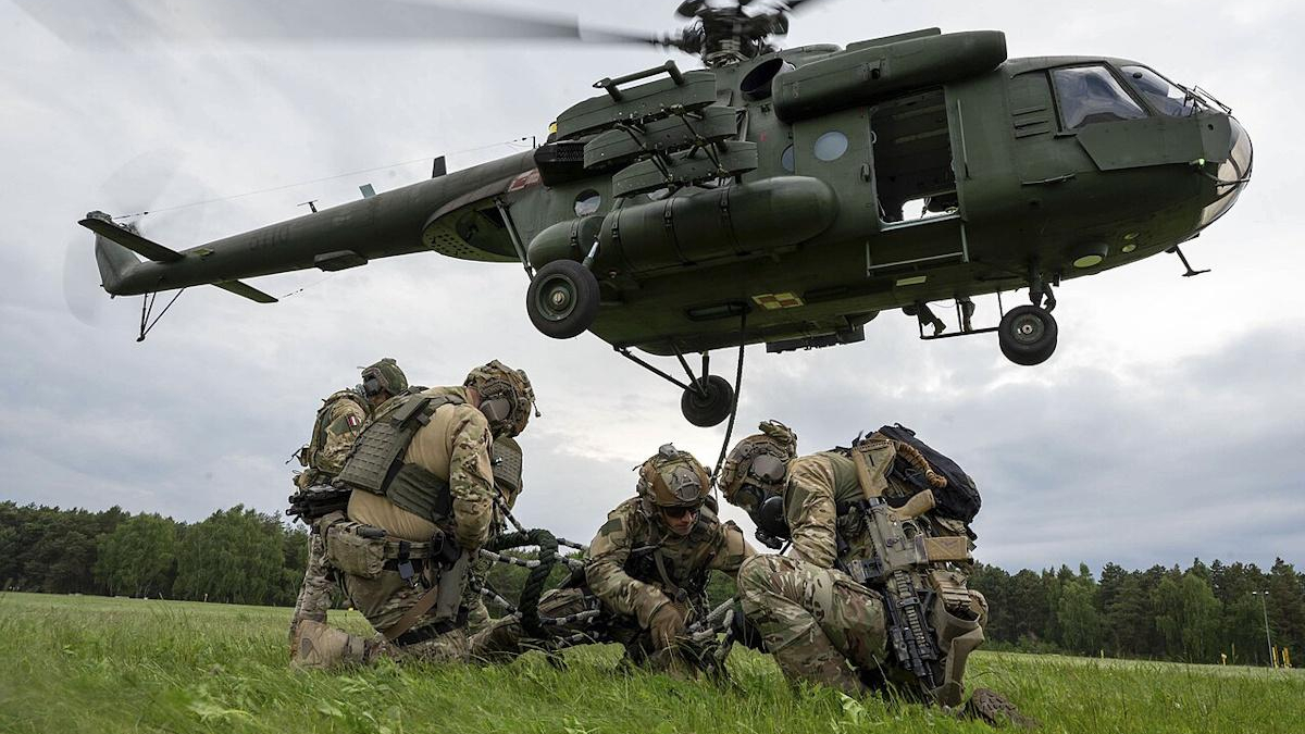 CH-47 Chinook helicopter hovering over soldiers in camouflage gear on a grassy field, preparing equipment.