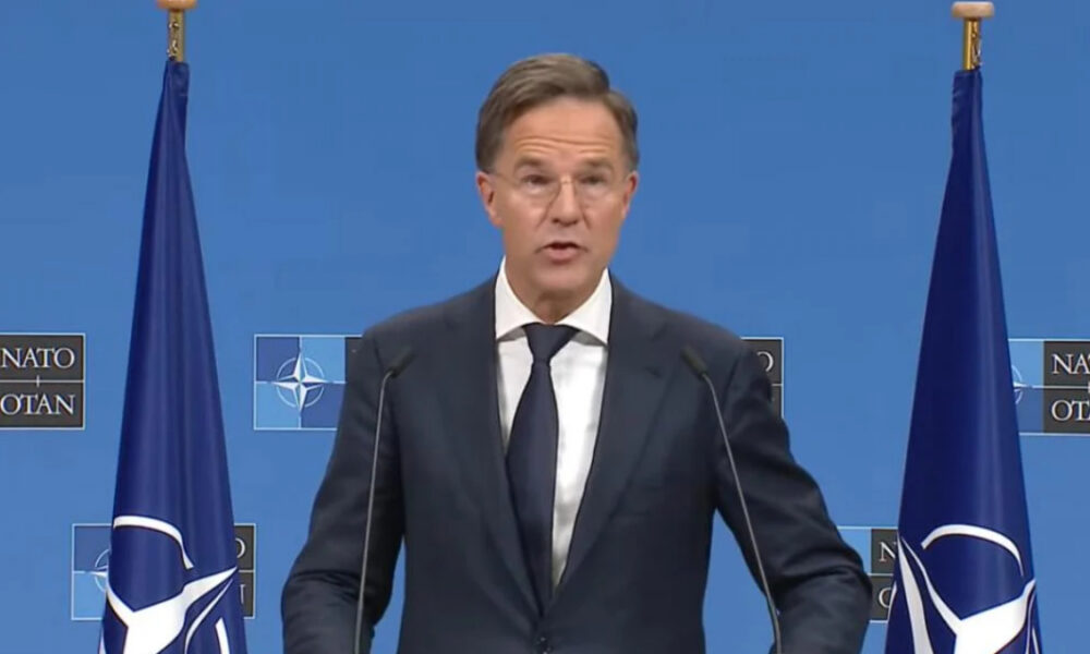 Middle-aged man in a dark suit and tie speaks at a NATO press briefing, with NATO flags on both sides and a blue backdrop behind him.