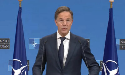Middle-aged man in a dark suit and tie speaks at a NATO press briefing, with NATO flags on both sides and a blue backdrop behind him.