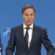 Middle-aged man in a dark suit and tie speaks at a NATO press briefing, with NATO flags on both sides and a blue backdrop behind him.