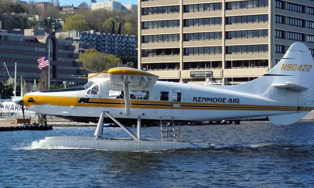 White Kenmore Air floatplane with yellow nose on a water runway, city buildings in the background.