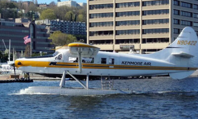 White Kenmore Air floatplane with yellow nose on a water runway, city buildings in the background.