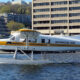 White Kenmore Air floatplane with yellow nose on a water runway, city buildings in the background.