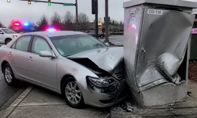 Silver sedan rear-ends a large metal structure at an intersection, with heavy front-end damage and deployed airbags visible through the windshield; emergency vehicles with flashing lights in the background.