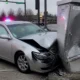 Silver sedan rear-ends a large metal structure at an intersection, with heavy front-end damage and deployed airbags visible through the windshield; emergency vehicles with flashing lights in the background.