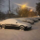 Nighttime street scene with a row of cars covered in fresh snow and glowing streetlights above.