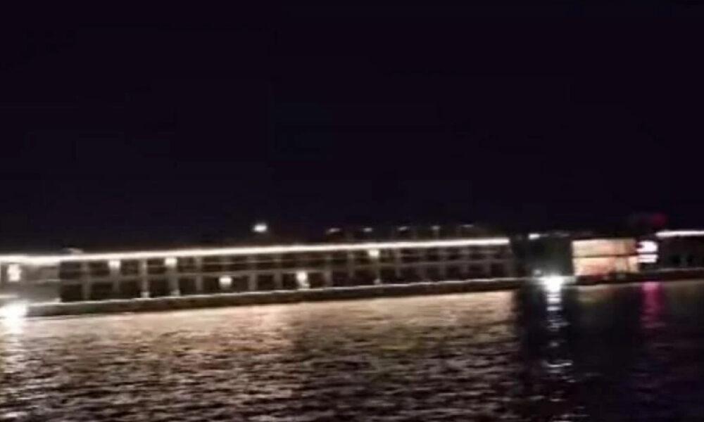 Nighttime view of a lit pier or boardwalk over calm water, with white lights along the railing and reflections on the surface.