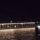 Nighttime view of a lit pier or boardwalk over calm water, with white lights along the railing and reflections on the surface.