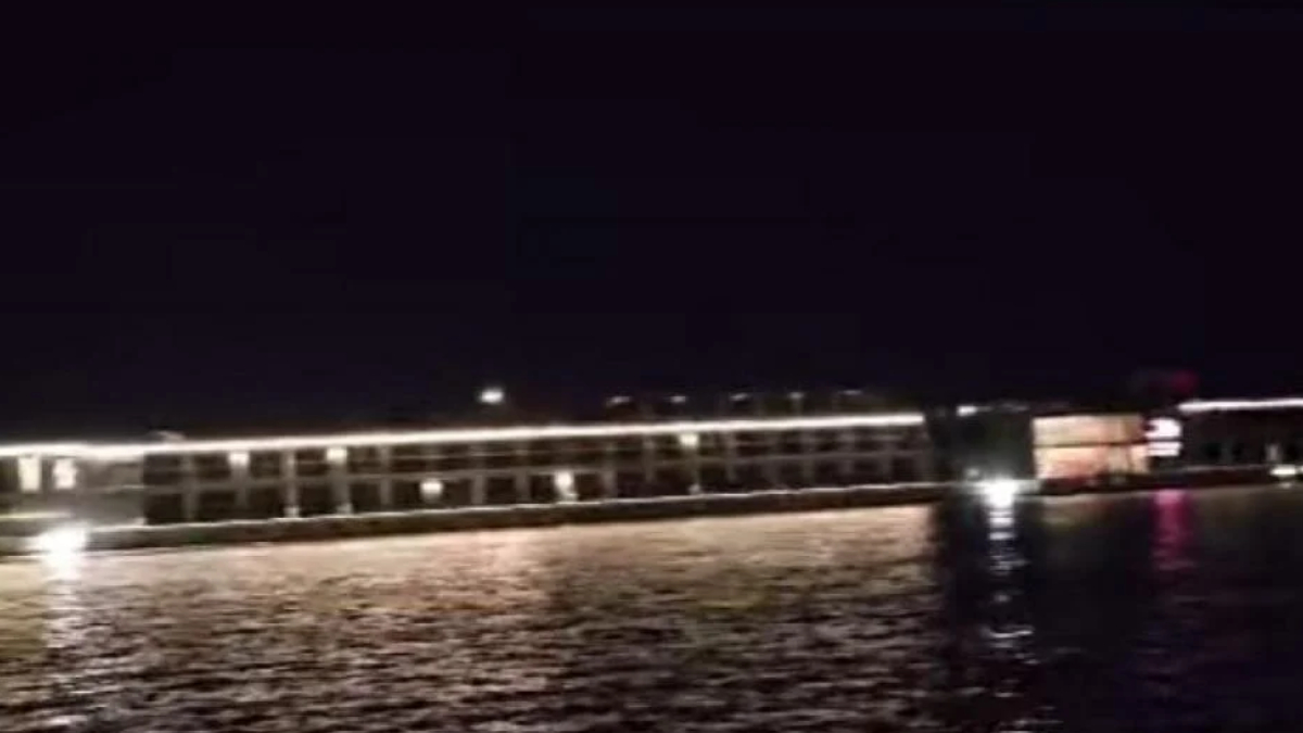 Nighttime view of a lit pier or boardwalk over calm water, with white lights along the railing and reflections on the surface.