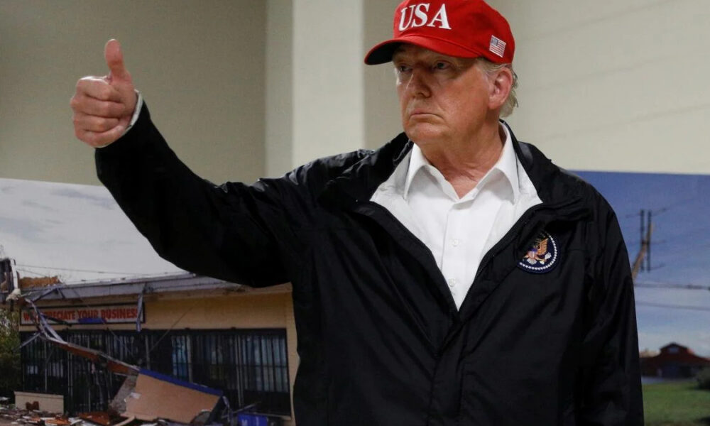 Man in a white shirt and black jacket gives a thumbs up, wearing a red USA cap indoors.
