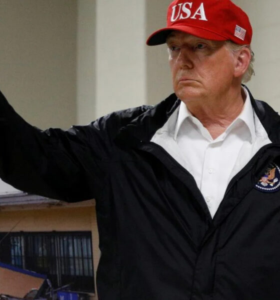 Man in a white shirt and black jacket gives a thumbs up, wearing a red USA cap indoors.