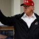 Man in a white shirt and black jacket gives a thumbs up, wearing a red USA cap indoors.