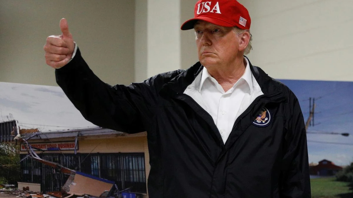 Man in a white shirt and black jacket gives a thumbs up, wearing a red USA cap indoors.