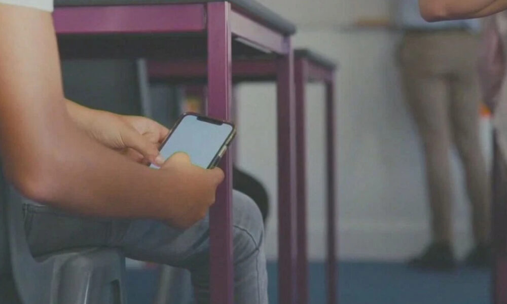 Student scrolling on a smartphone while seated in a classroom, purple chair legs visible nearby.