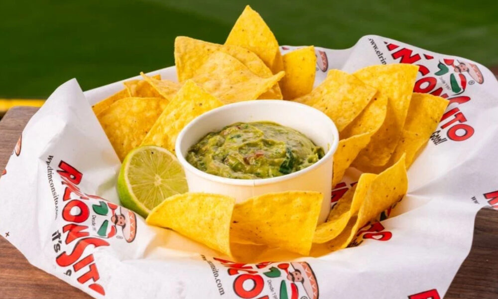 Tortilla chips arranged around a white cup of chunky guacamole with a lime wedge, served on a parchment liner outdoors on a wooden table.