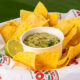 Tortilla chips arranged around a white cup of chunky guacamole with a lime wedge, served on a parchment liner outdoors on a wooden table.